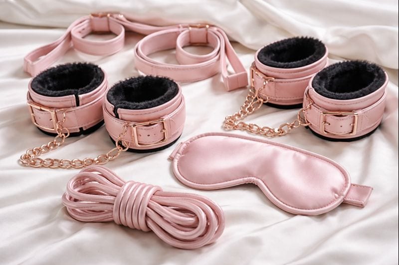 A set of pink faux leather restraints, including wrist and ankle cuffs with gold chains, a blindfold, and a length of soft rope-perfect beginner BDSM kits that are safe and fun, arranged on a white satin sheet.