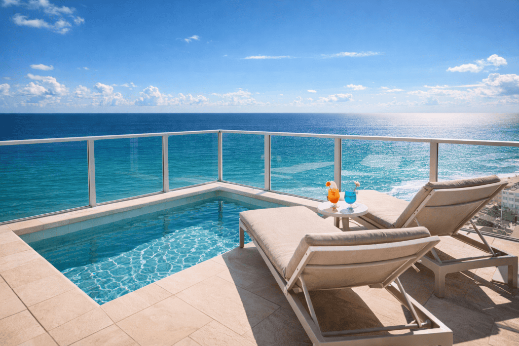 Two lounge chairs and a small table with drinks sit beside a private pool on a balcony overlooking the ocean under a clear blue sky at one of the premier swinger hotels near Miami.