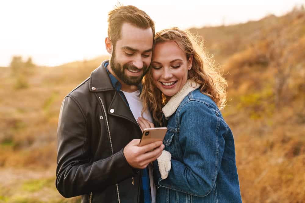 A man and woman stand outdoors, smiling as they look at a smartphone together. Dressed in jackets under the sun, they enjoy nature-perhaps reviewing lifestyle profile advice to get more matches swinger style.