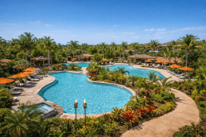 Large outdoor resort pool surrounded by tropical plants, lounge chairs with orange umbrellas, and several cabanas under a clear blue sky at one of the top swinger resorts near Tampa.
