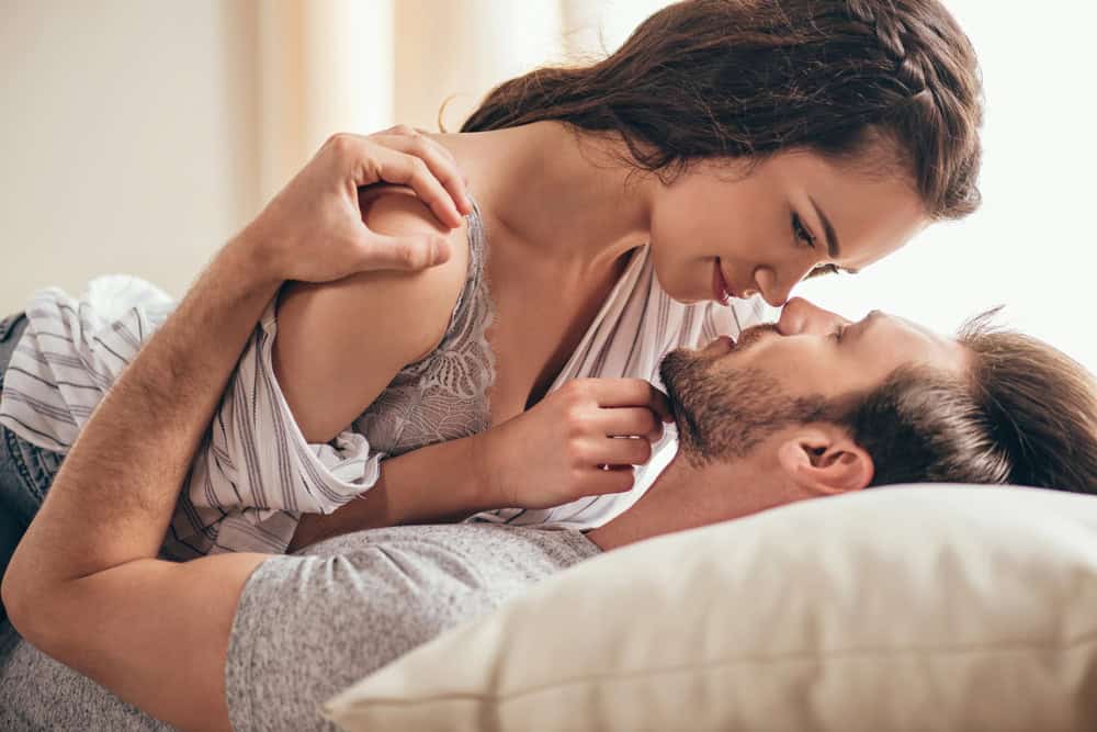 A woman lies on top of a man on a bed, both looking into each other's eyes closely, exploring the excitement of the swingers lifestyle.